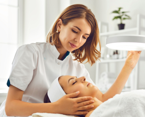 Cosmetologist Directing Lamp To Woman For Facial Treatment